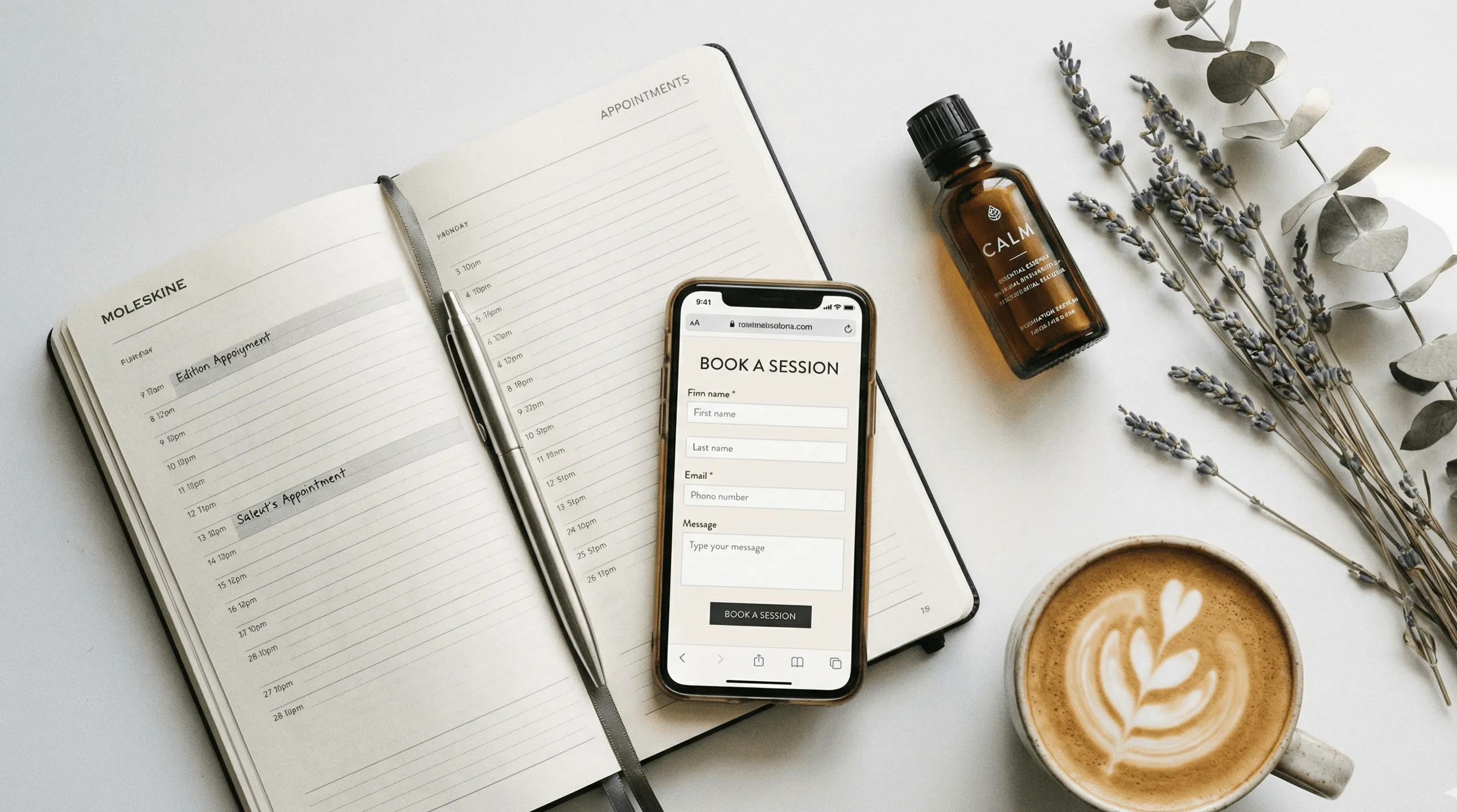 Open Moleskine planner with handwritten appointments, a smartphone showing a booking form, a bottle of Calm essential essence, lavender sprigs, and a cup of latte with leaf latte art on a white surface.