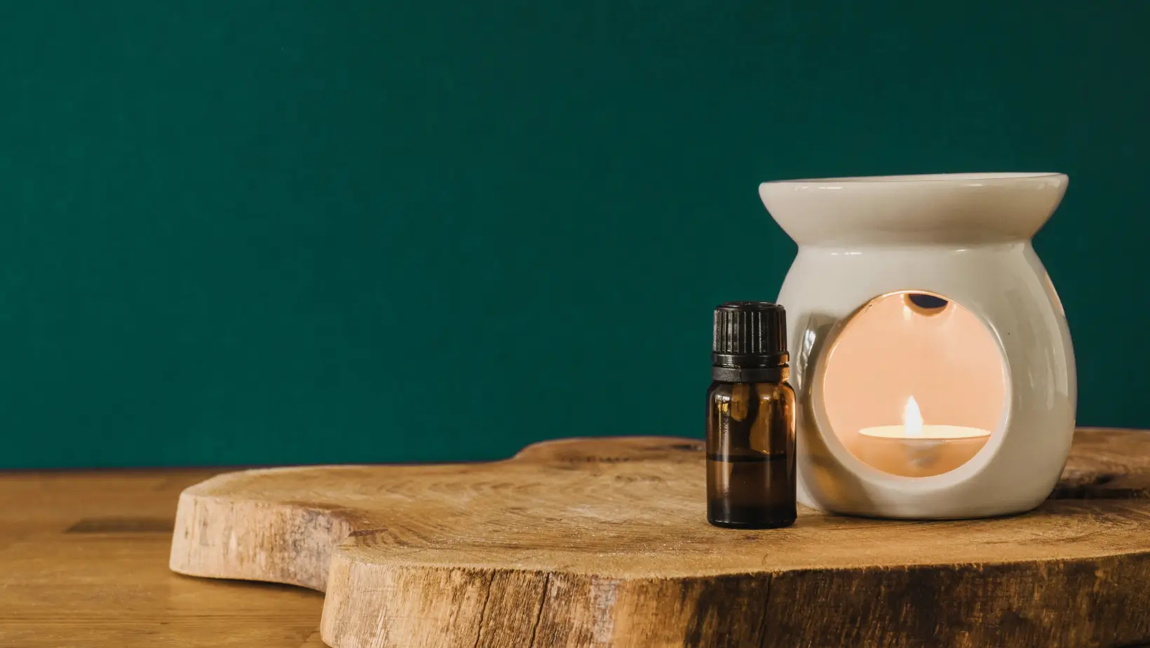 Small amber essential oil bottle next to a lit white ceramic oil burner on wooden surface.