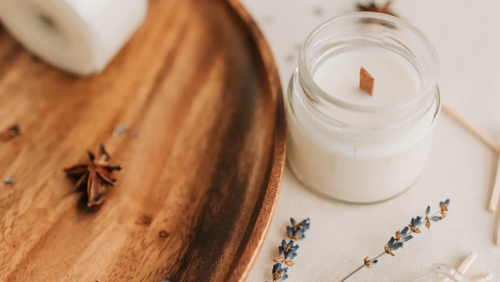White candle with wooden wick in a glass jar beside dried lavender and a wooden plate.
