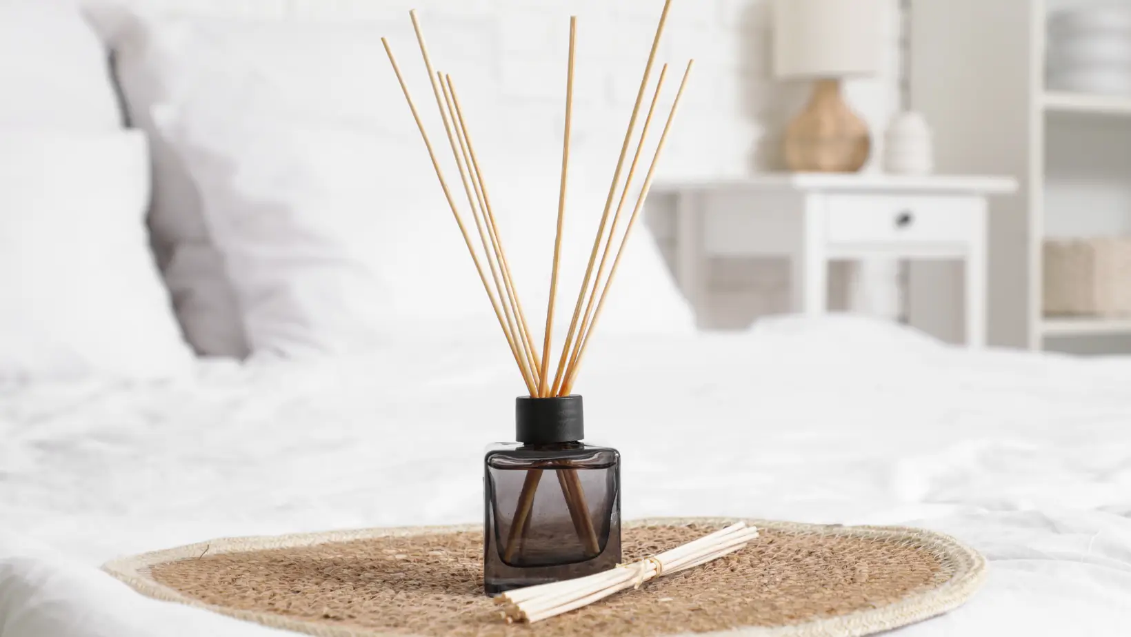 A glass diffuser bottle with wooden reed sticks on a woven mat in a white bedroom setting.