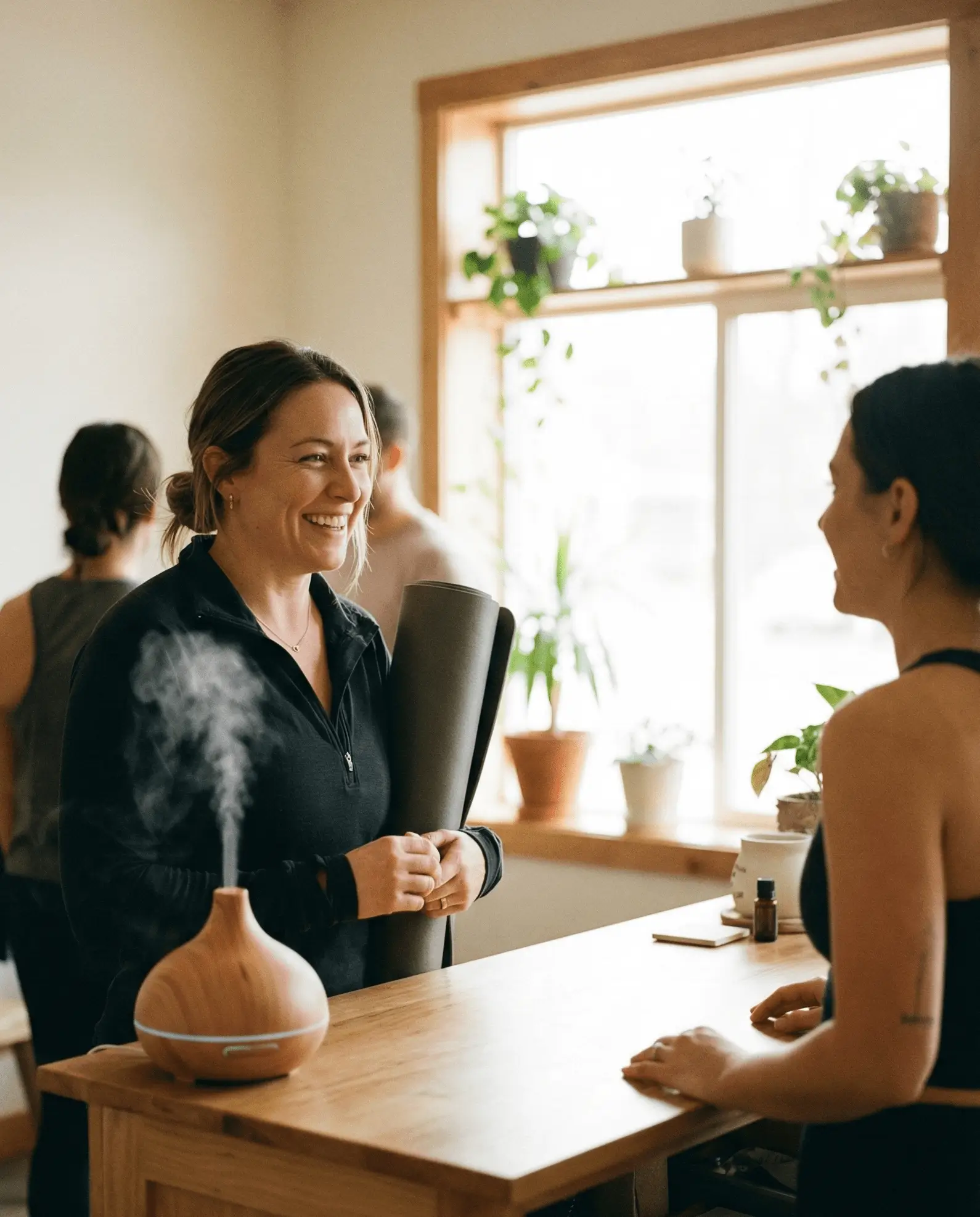 Two women smiling and talking across a wooden counter in a bright room with potted plants and a diffusing humidifier.