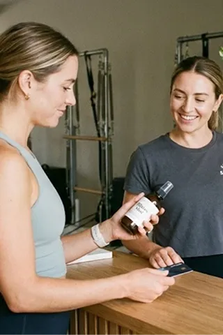 Two women standing at a wooden counter, one holding a bottle of skincare product and the other smiling.
