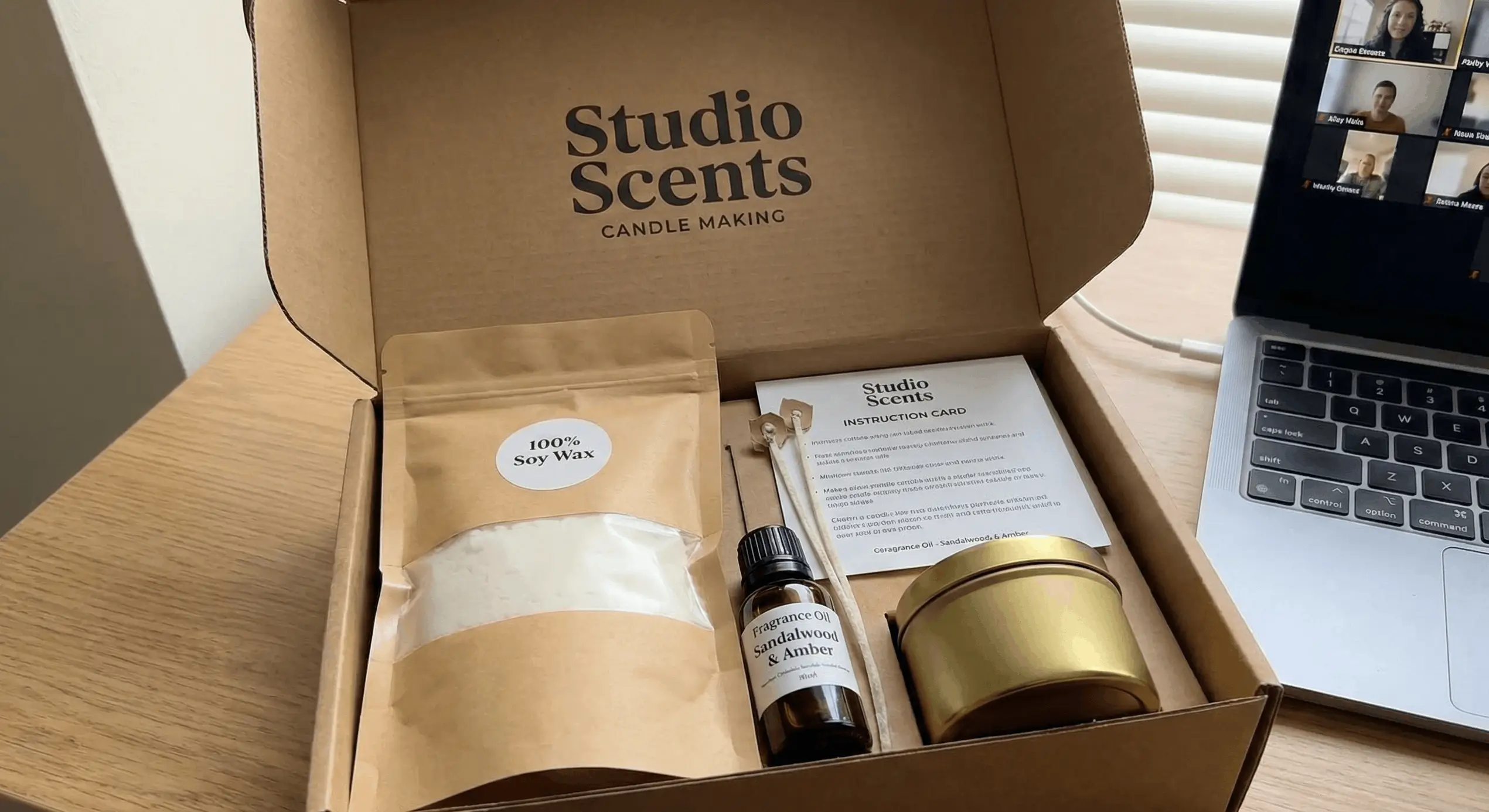 Open Studio Scents candle making kit with 100% soy wax, sandalwood and amber fragrance oil, metal tin, wick holder, and instruction card on a wooden table beside a laptop.