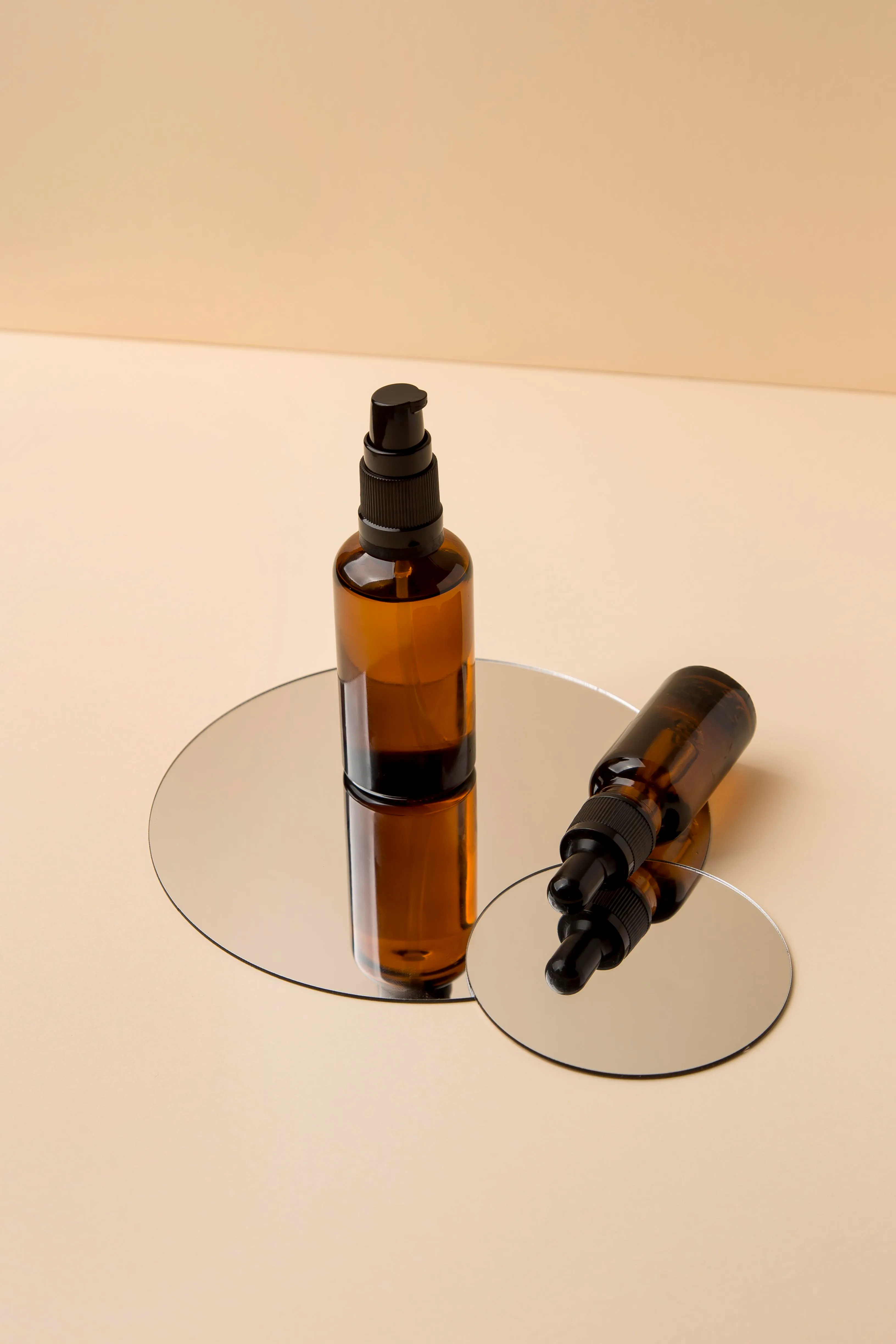 Two amber glass dropper bottles, one standing and one lying on its side, placed on round mirrors against a beige background.
