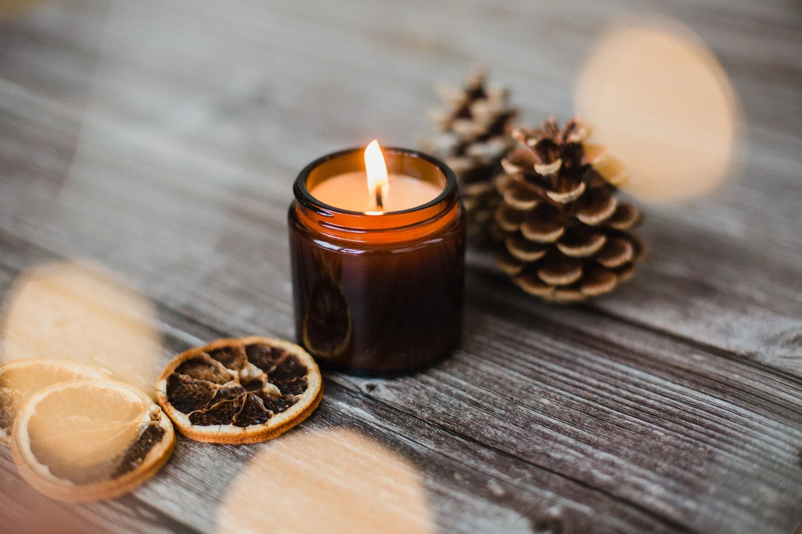 Lit candle in a brown glass jar on a wooden surface surrounded by pine cones and dried citrus slices.