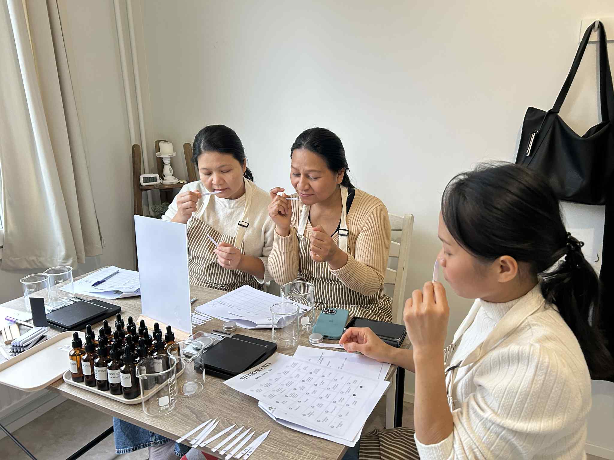 Three women sitting at a table smelling fragrance strips, surrounded by bottles, beakers, and scent evaluation sheets.