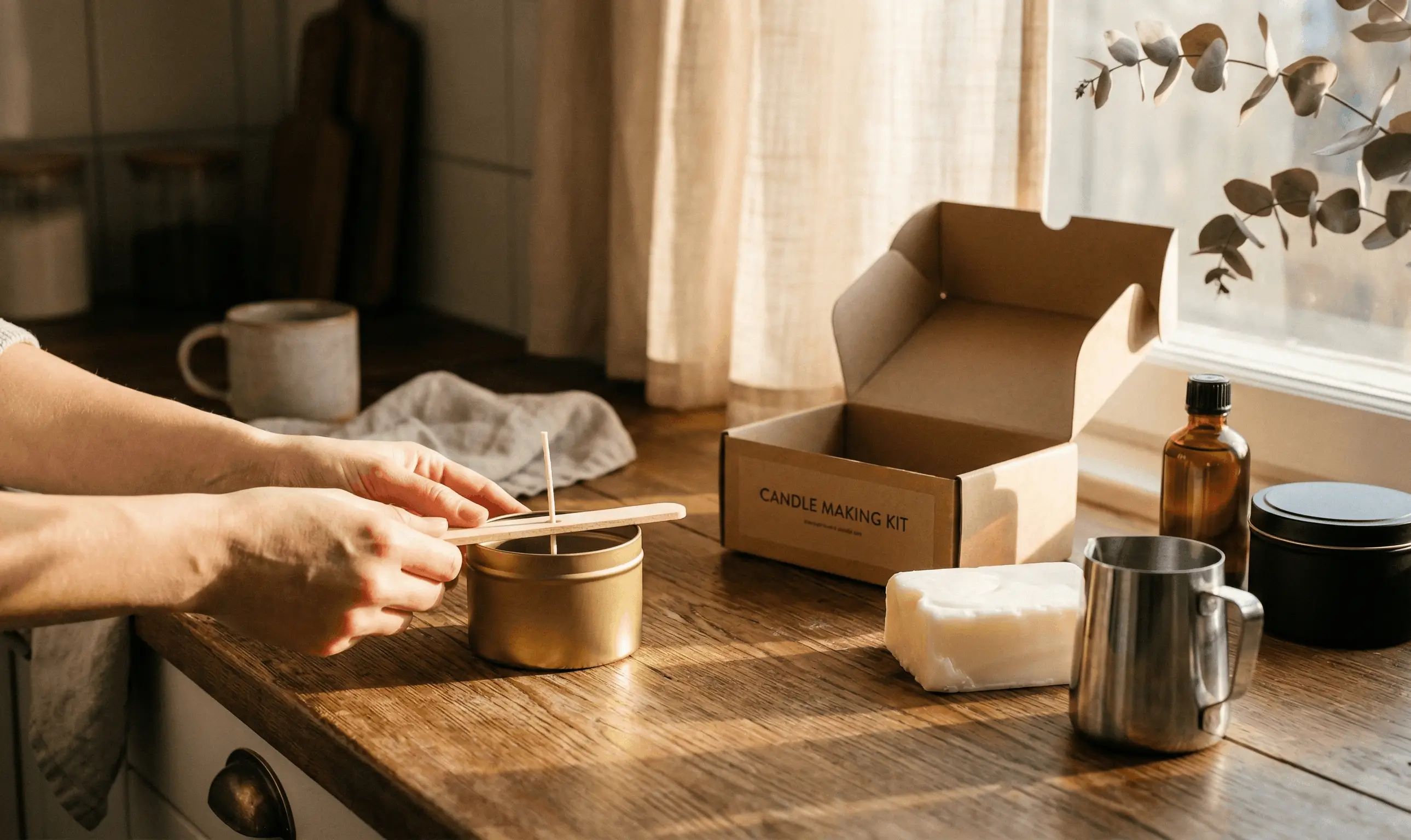 Person placing a wick into a metal candle container on a wooden countertop with a candle making kit box and supplies nearby.