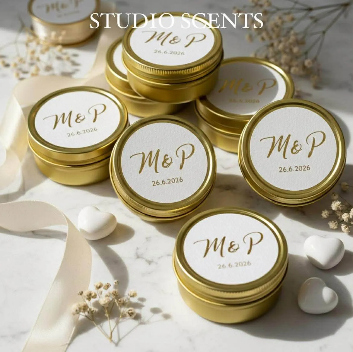 Gold tins with white lids labeled 'M&P 26.6.2026' arranged on a marble surface with cream ribbon, dried flowers, and white heart-shaped objects.