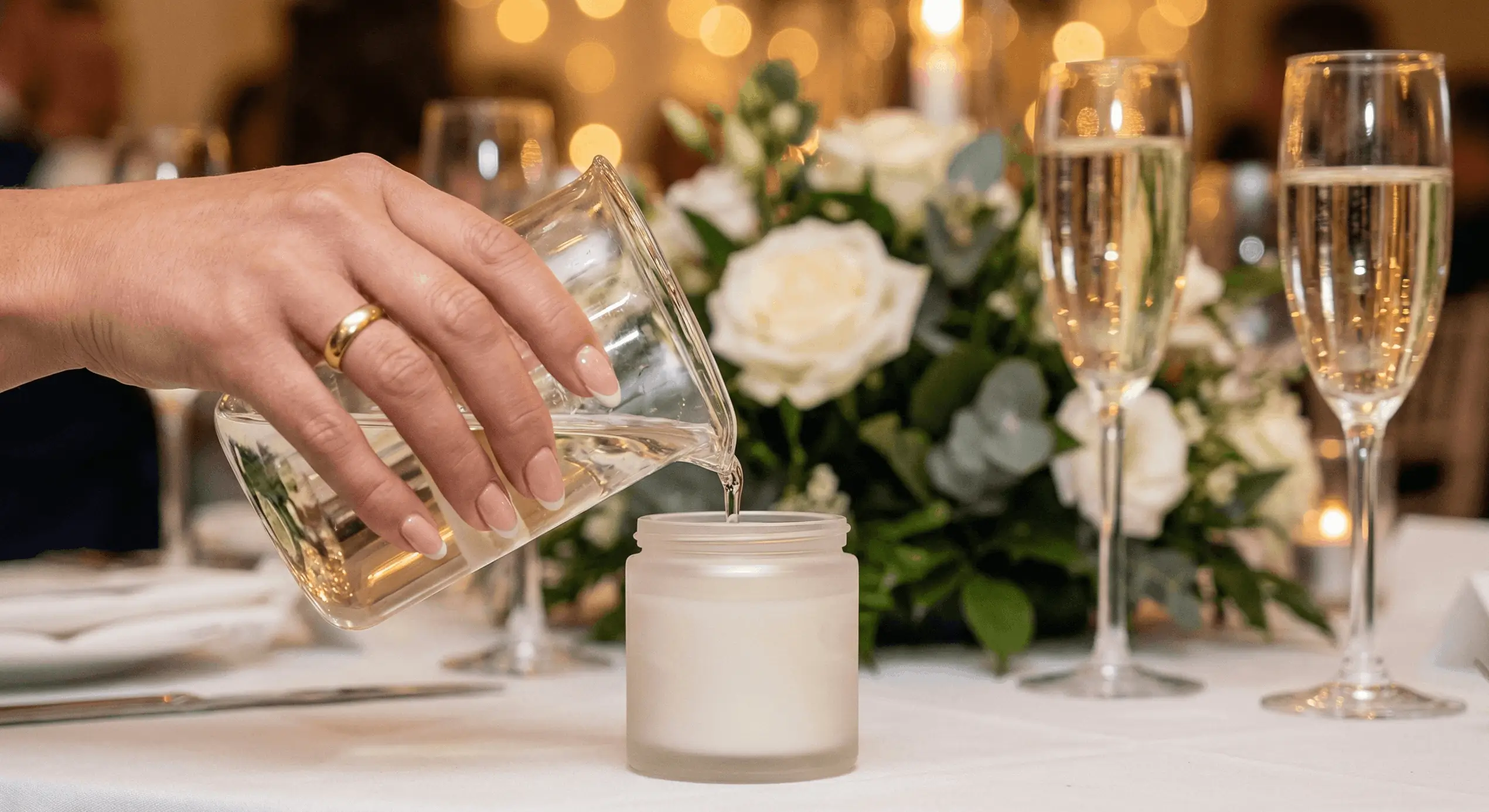 Hand with manicured nails and a gold ring pouring liquid from a glass pitcher into a frosted glass candle holder on a decorated table with white roses and champagne glasses.