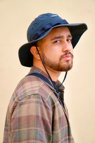 Man with beard wearing a black bucket hat and plaid shirt looking sideways against a beige background.