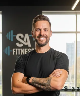 Smiling man with short hair and tattoo on forearm wearing a black t-shirt, standing with arms crossed in a gym setting.