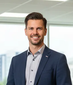 Smiling man with brown hair wearing a dark blue blazer and light gray shirt, standing in a modern office setting with large windows.