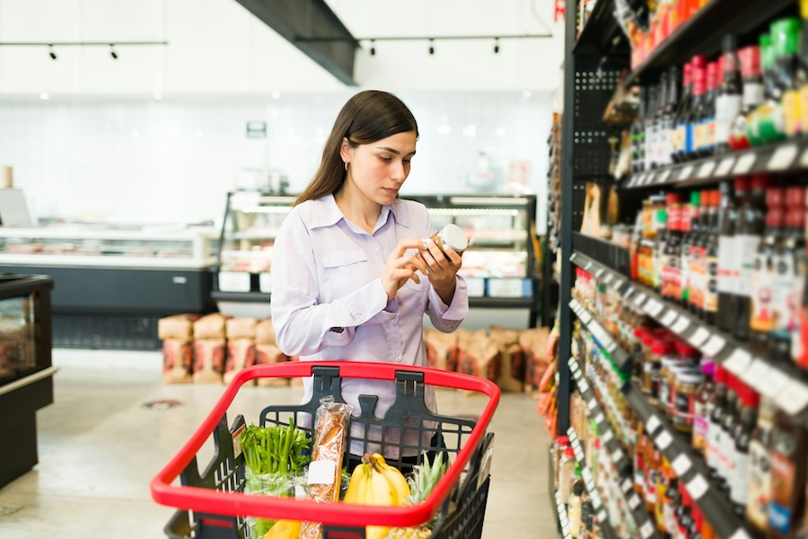 Woman reading the label of a product while standing next to shelves in a grocery store with a shopping cart containing bananas, baguette, and green vegetables.