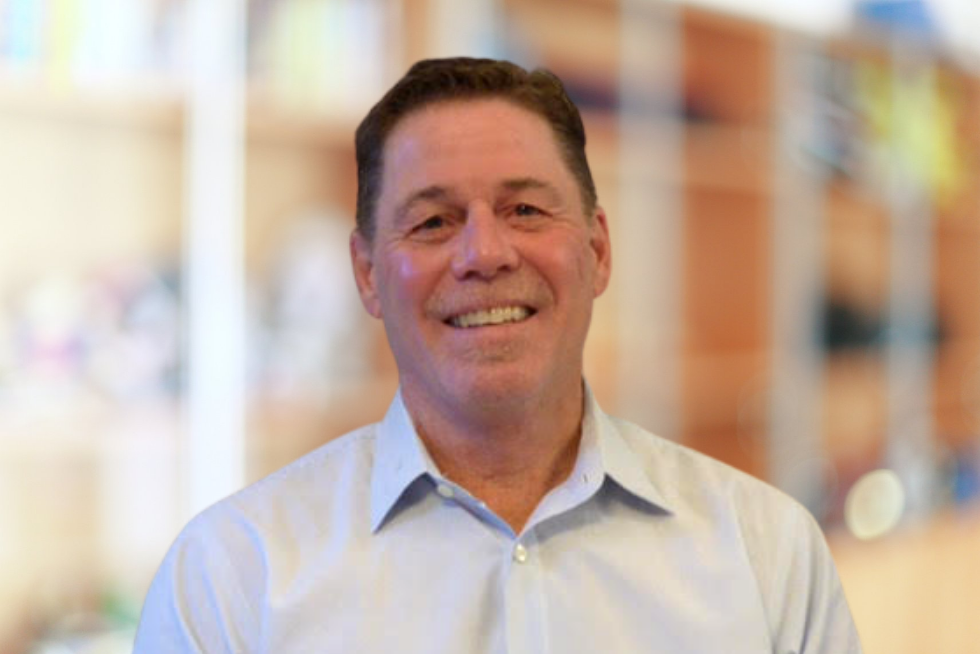 Smiling middle-aged man in a light blue collared shirt with a blurred indoor background.