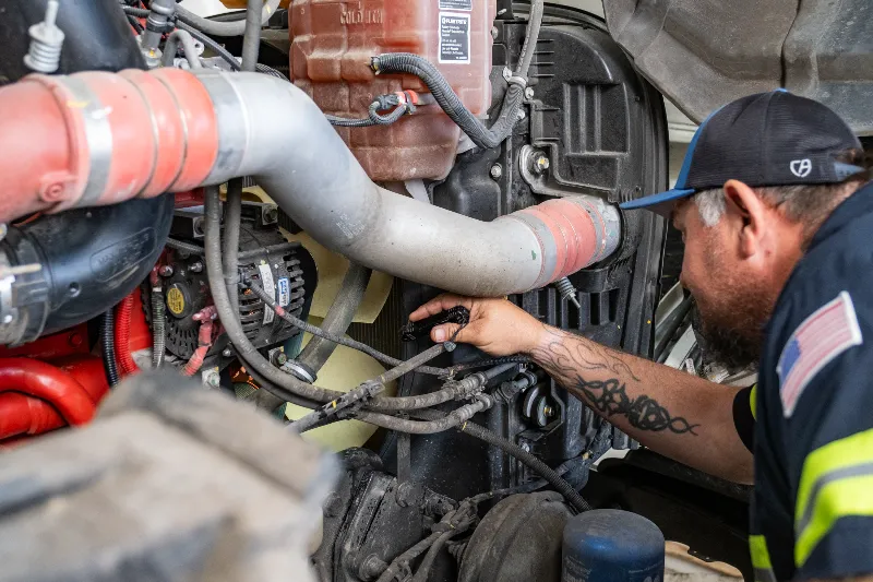 Summer maintenance inspection on a heavy-duty truck engine, with a technician checking hoses and cooling system components.