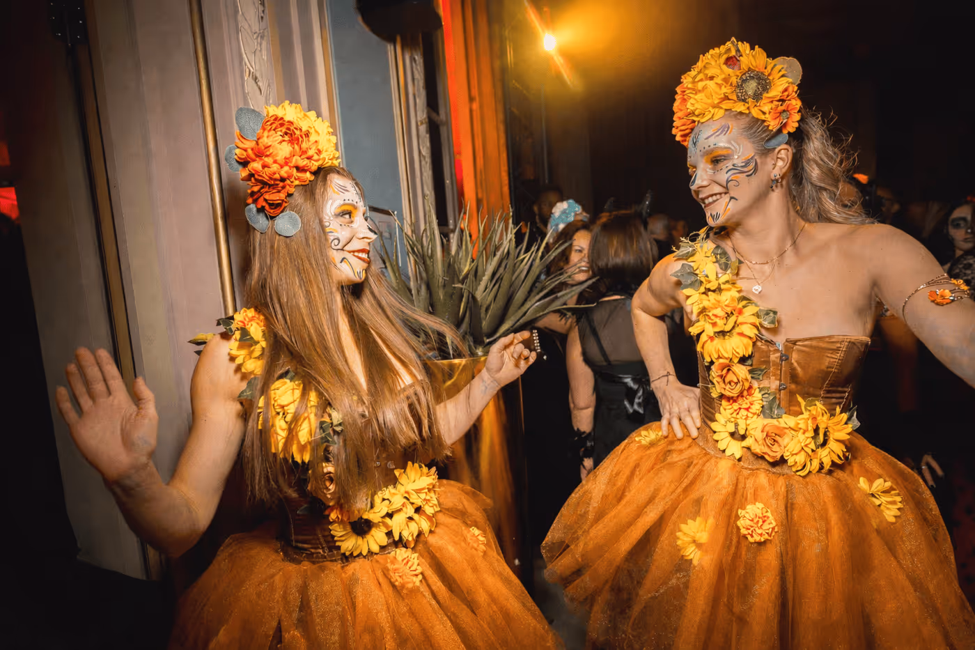 Deux femmes souriantes en costumes orange avec décorations florales jaunes et maquillage artistique au visage lors d'une fête.