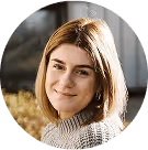 Frau mit schulterlangem Haar lächelt in die Kamera, trägt einen grauen Pullover, im Hintergrund sonnige Natur.