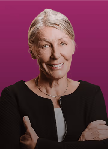 Ältere Frau mit grauem Haar und verschränkten Armen, lächelnd vor violettem Hintergrund.