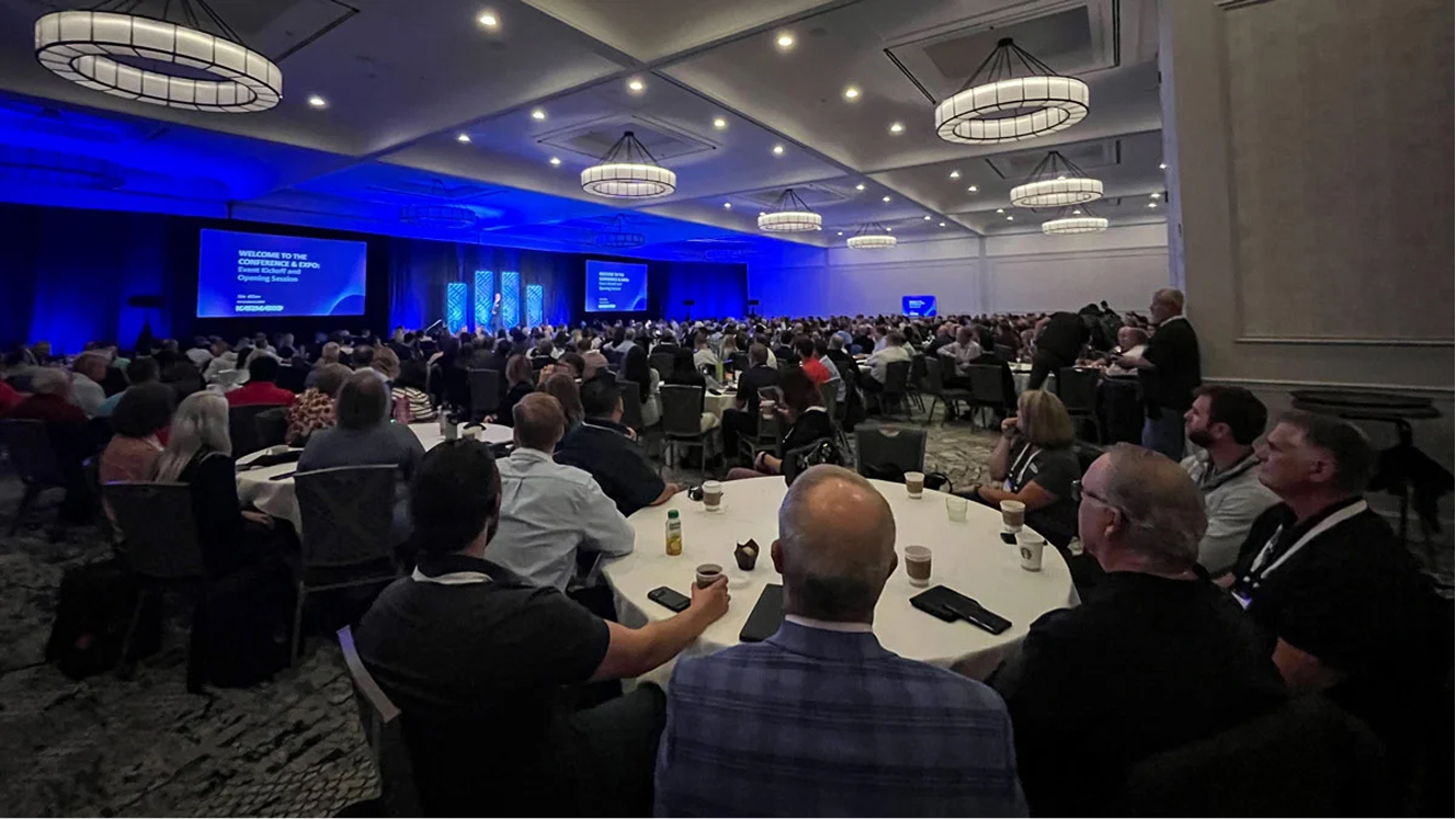 Large audience seated at round tables in a conference room watching a presenter on stage with blue lit backdrop and screens.