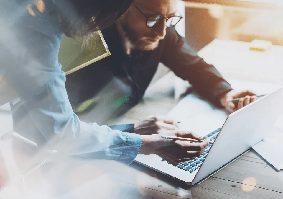 Two people collaborating over a laptop on a wooden table in a warmly lit office.