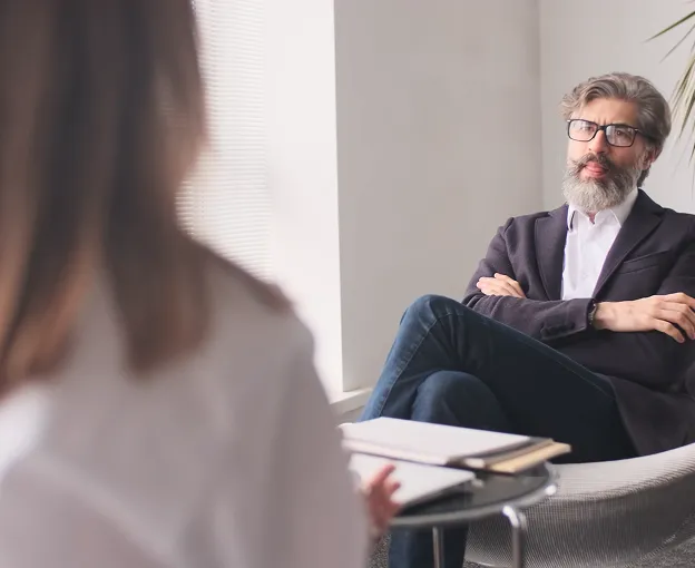 Man with glasses and gray beard sitting in a chair with arms crossed, listening to a woman speaking in an office setting.