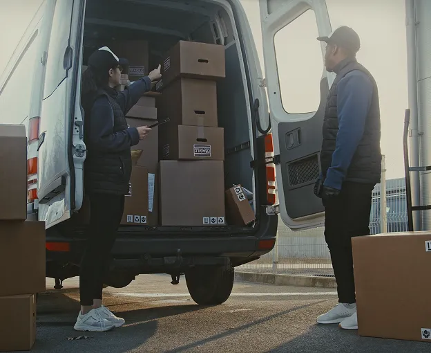 Two delivery workers loading cardboard boxes into the back of a white delivery van in an outdoor setting.