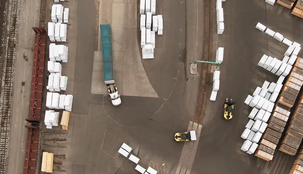 Aerial view of a shipping yard with a white truck, two yellow forklifts moving pallets, and stacks of white boxes on pavement near train tracks.