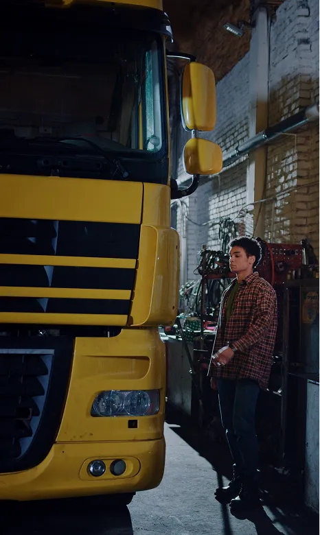 Young man in a plaid shirt standing next to the front side of a large yellow truck inside a dimly lit garage.
