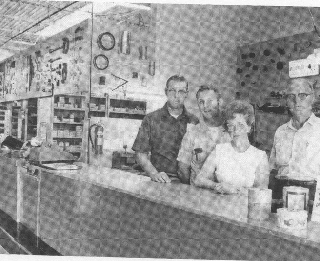 Black-and-white photo of three men and one woman standing behind a long counter in a hardware or parts store with shelves and display panels in the background.