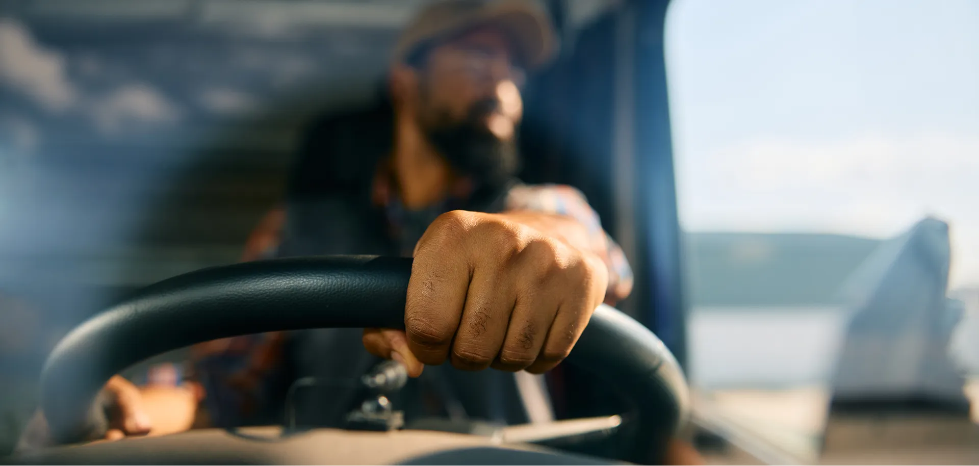 Close-up of a man's hand gripping a steering wheel with a blurred view of the man in the background inside a vehicle.