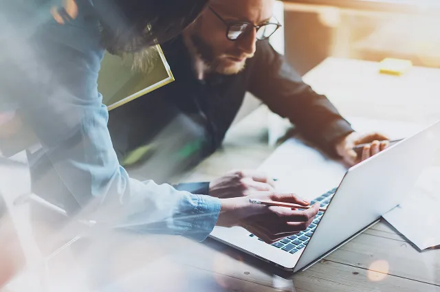 Two people collaborating while working on a laptop at a wooden desk, one pointing at the screen with a pen.