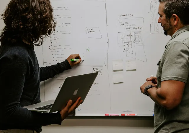 Two people collaborating in front of a whiteboard with notes and diagrams while one holds a laptop and writes with a marker.