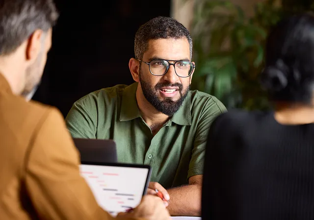 Man with glasses and beard in green shirt talking to two people, one holding a laptop.