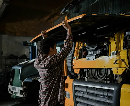 Man in plaid shirt inspecting the raised hood of a yellow truck in a dimly lit garage.