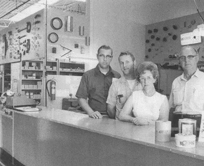 Black and white photo of two men and one woman standing behind a store counter with another man standing beside them in a workshop or hardware store.