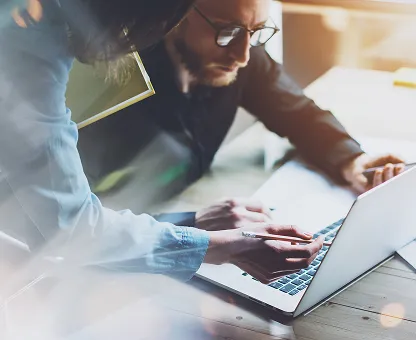 Two people collaborating over a laptop, one pointing at the keyboard with a pen.