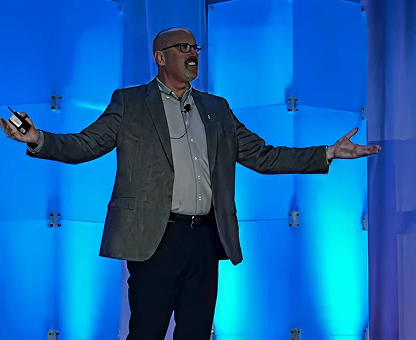 Man with glasses speaking on stage with blue-lit background, holding a remote and gesturing with open arms.