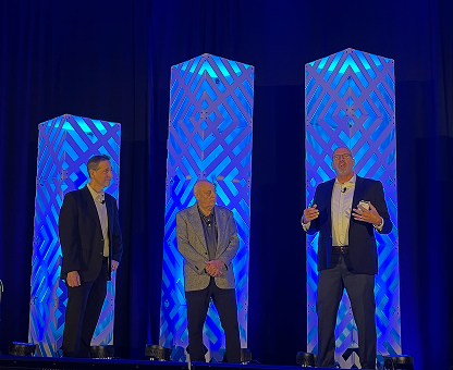 Three men standing on stage with blue illuminated geometric-patterned columns behind them.