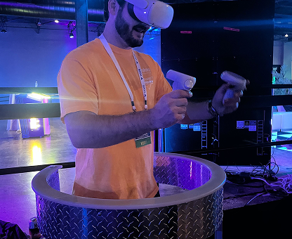 Man wearing a virtual reality headset and holding VR controllers standing inside a circular metal platform in a dimly lit room with blue lighting.