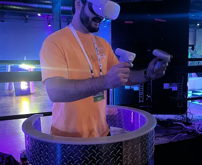 Man wearing a virtual reality headset and holding VR controllers standing inside a circular metal platform in a dimly lit room with blue lighting.