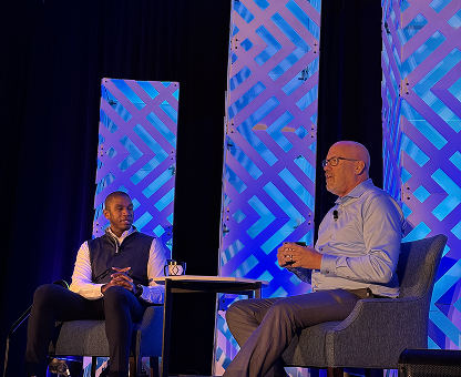 Two men seated and engaged in a discussion on stage with geometric blue and purple panels in the background.