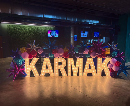 Illuminated letters spelling 'KARMAK' surrounded by colorful balloons and star-shaped decorations in a dimly lit indoor space.