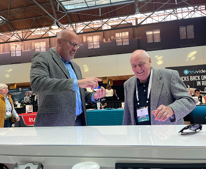 Two smiling men in blazers at a trade show booth, one holding a small object while the other watches.