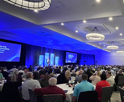 Large audience seated at round tables watching a speaker present on stage with blue-lit screens in a conference room.