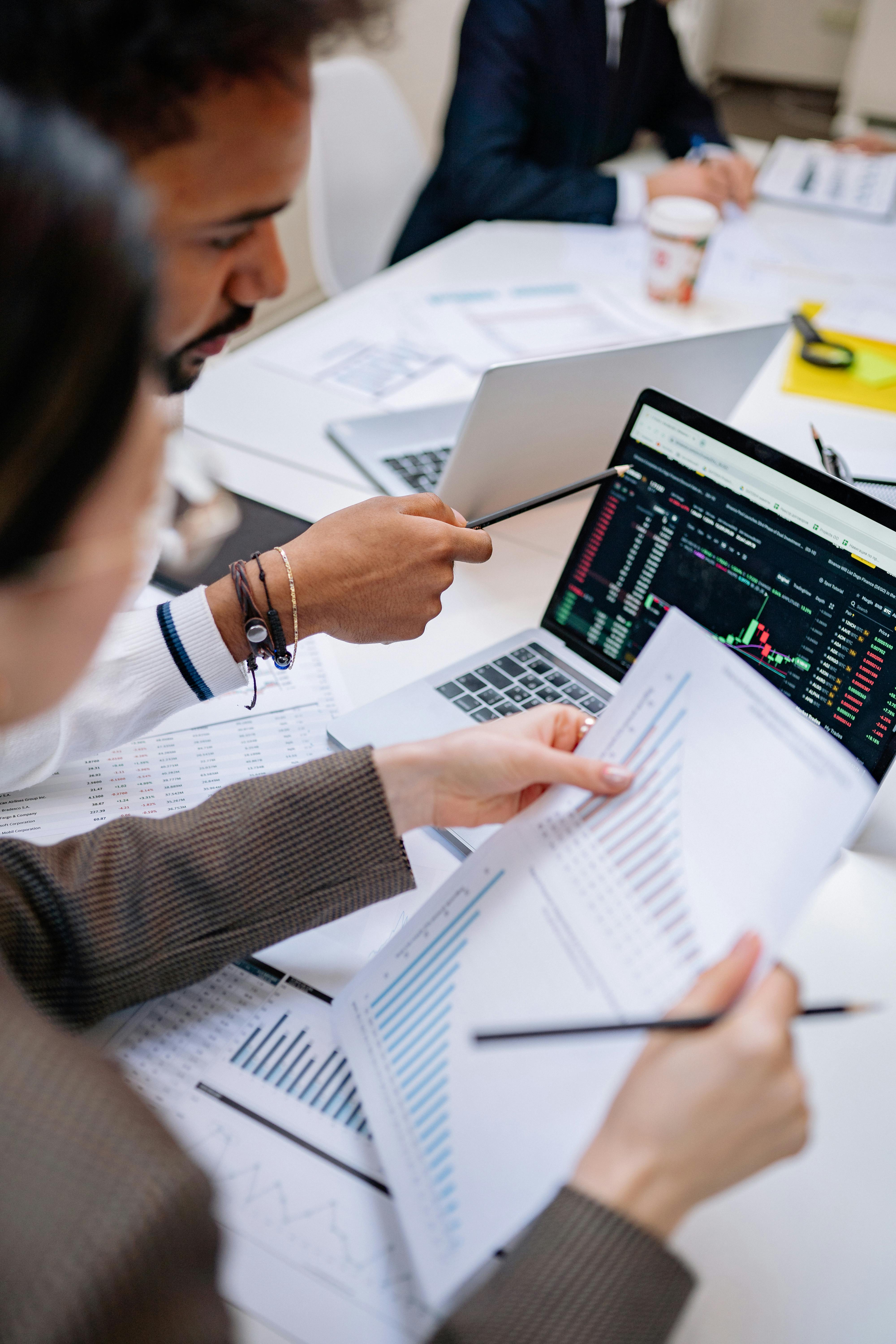 Two people analyzing financial charts and stock market data on a laptop and printed documents at a white table.