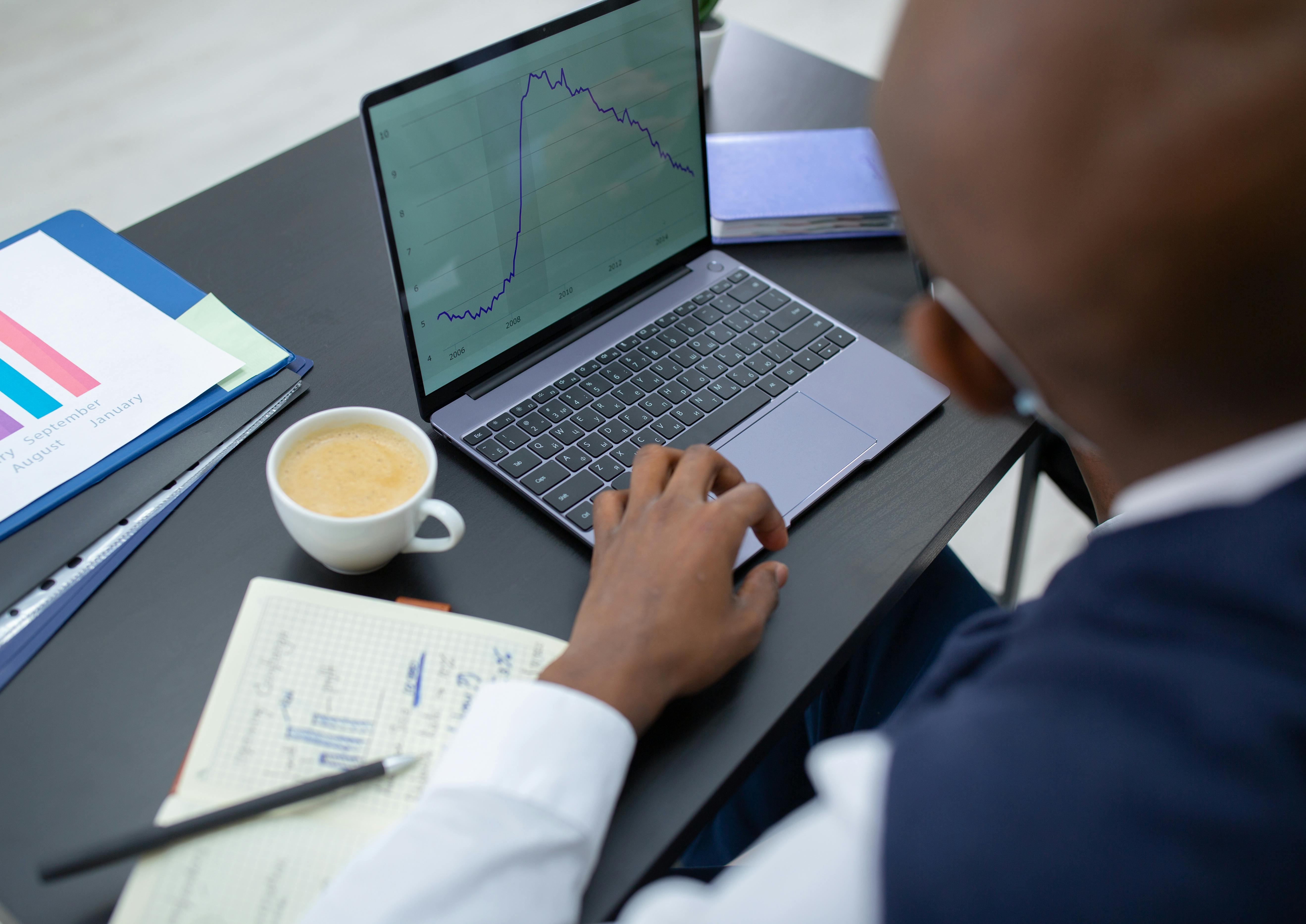 Person working on a laptop with financial charts displayed on the screen, beside a cup of coffee and a notebook with handwritten notes on a desk.