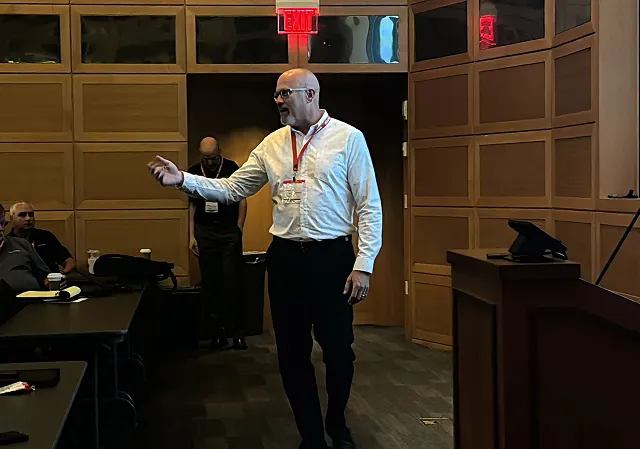 Man in white shirt and glasses speaking in a conference room with wooden walls and an exit sign above the door.