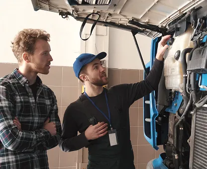 Mechanic explaining engine parts to a customer beside an open truck hood.