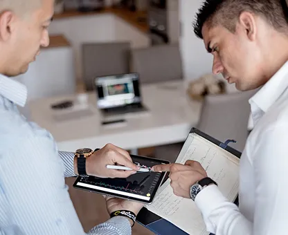 Two men analyzing financial charts, one using a tablet and the other pointing at a paper notebook.