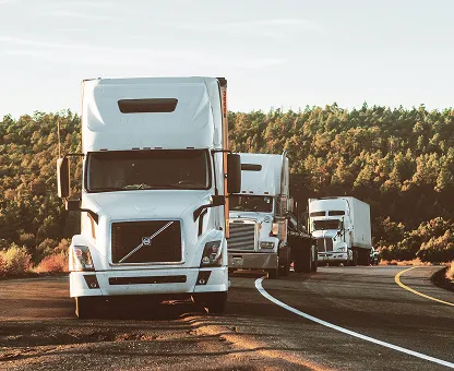 Three white semi-trailer trucks driving on a curved road with a forest in the background.